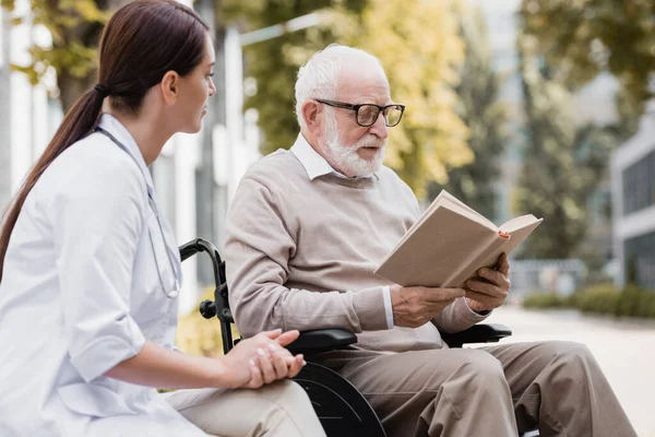 Handicapé dans les lunettes de lecture livre près travailleur social à l'extérieur — Photo de stock