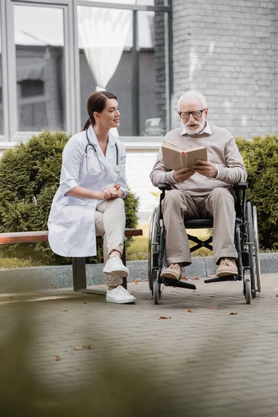 Handicapped aged man reading book in wheelchair near social worker sitting on bench on blurred foreground — Stock Photo