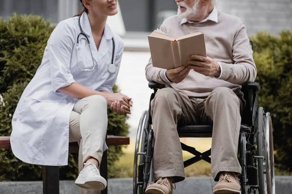 Cropped view of social worker on bench near handicapped man reading book on wheelchair outdoors — Stock Photo