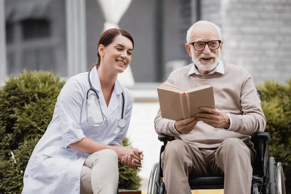 Cheerful geriatric nurse near aged disabled man reading book in wheelchair outdoors — Stock Photo