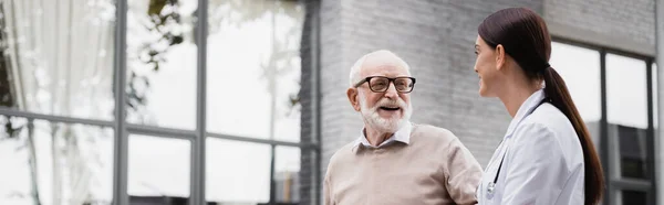 Alegre anciano y trabajador social mirándose mientras pasean afuera, pancarta - foto de stock