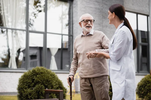 Enfermera geriátrica que apoya a un hombre anciano paseando con bastón - foto de stock