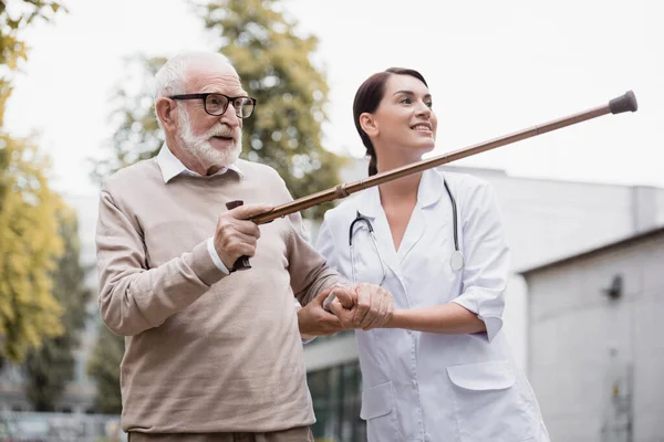 Aged man pointing with walking stick away near smiling geriatric nurse — Stock Photo
