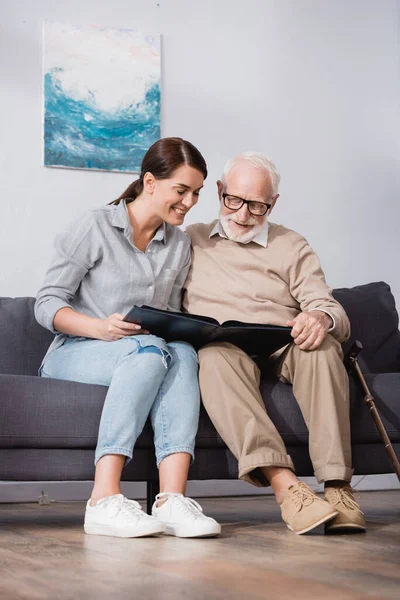 Fröhlicher Vater und Tochter blättern im Fotoalbum, während sie zu Hause auf dem Sofa sitzen — Stockfoto