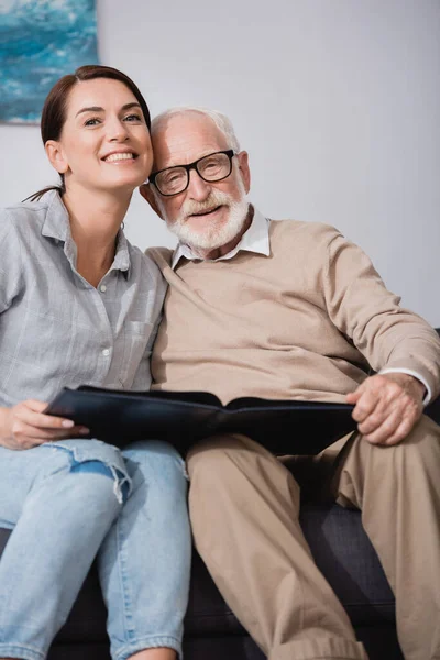 Happy father with adult daughter smiling at camera while holding photo album — Stock Photo