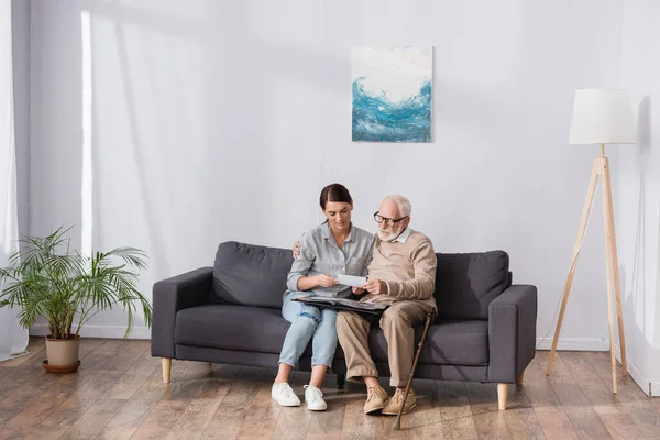 Älterer Vater mit erwachsener Tochter blättert im Fotoalbum, während er zu Hause auf dem Sofa sitzt — Stockfoto