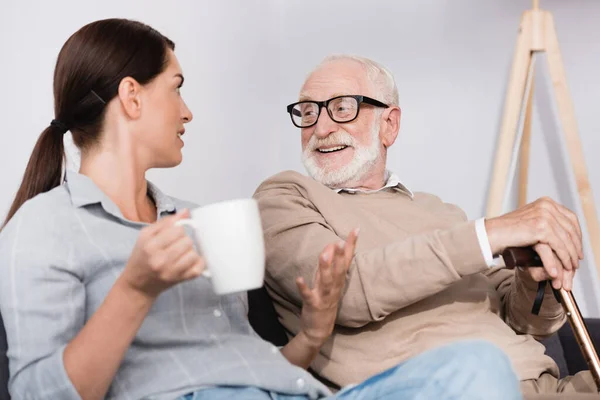 Brunette woman holding tea and gesturing while talking to cheerful elderly father — Stock Photo