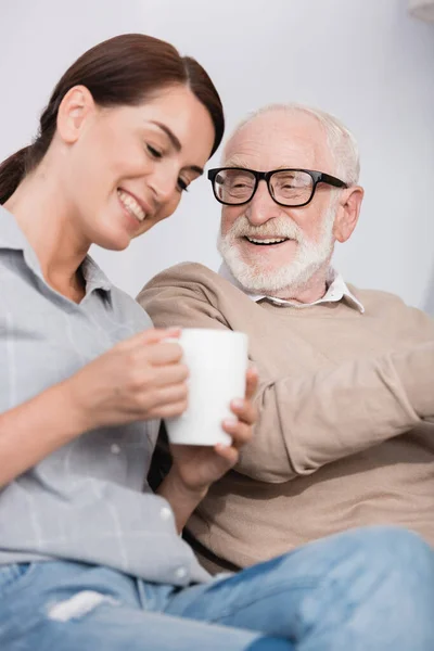 Donna allegra con tazza di tè che parla con padre sorridente invecchiato in primo piano sfocato — Foto stock