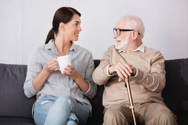 Donna bruna con tazza di tè e sorridente uomo anziano con bastone da passeggio che parla mentre siede sul divano a casa — Foto stock