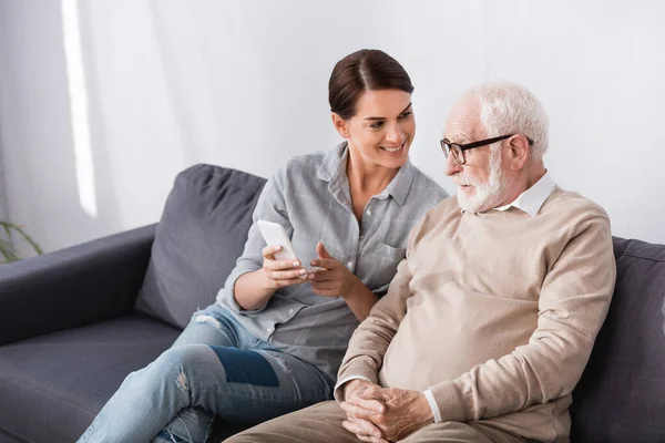 Cheerful woman pointing with hand at mobile phone near confused aged dad — Stock Photo