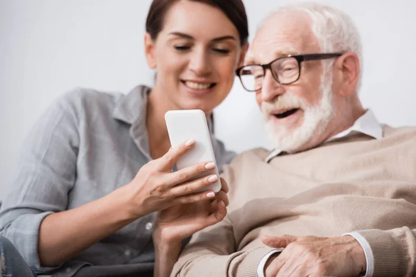 Sonriente mujer mostrando el teléfono móvil a alegre padre anciano sobre fondo borroso - foto de stock