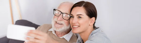 Happy woman taking selfie on smartphone with cheerful aged father on blurred foreground, banner — Stock Photo