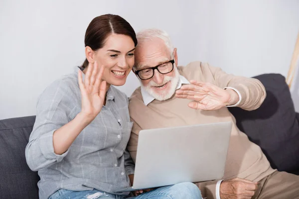 Alegre anciano hombre con hija saludando las manos mientras tiene video chat en el ordenador portátil - foto de stock