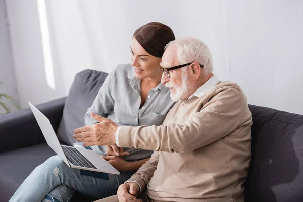 Пожилой мужчина указывает рукой на ноутбук рядом с взрослой дочерью, сидя на диване дома — стоковое фото