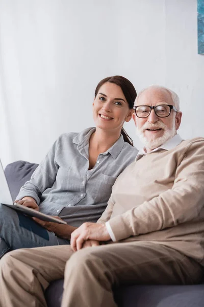 Happy woman with laptop and aged father looking at camera on blurred foreground — Stock Photo