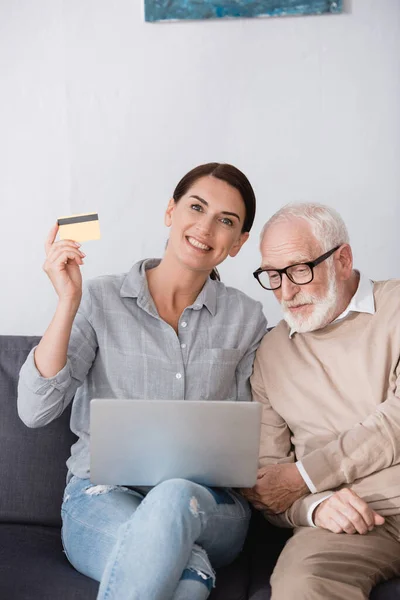 Mujer alegre sosteniendo el ordenador portátil y la tarjeta de crédito mientras está sentado cerca de padre anciano - foto de stock