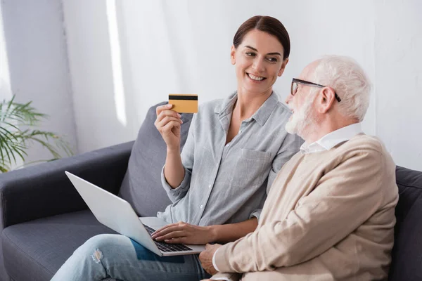 Улыбающаяся женщина с кредитной картой и ноутбуком рядом с отцом в возрасте, сидя на диване дома — стоковое фото