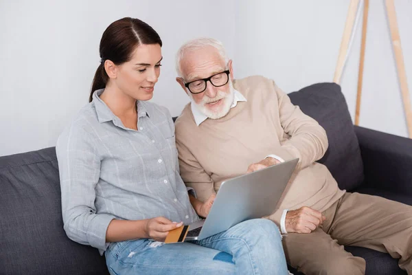 Mulher morena segurando cartão de crédito ao usar laptop junto com pai idoso — Fotografia de Stock