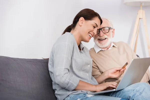 Веселая женщина с ноутбуком и кредитной картой рядом с счастливым пожилым отцом — стоковое фото
