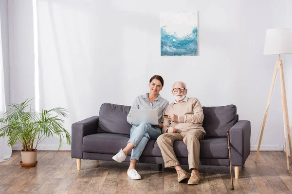 Anciano con hija adulta sentado en el sofá en casa con ordenador portátil y tarjeta de crédito - foto de stock