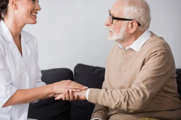 Side view of aged man in eyeglasses holding hands with social worker at home — Stock Photo