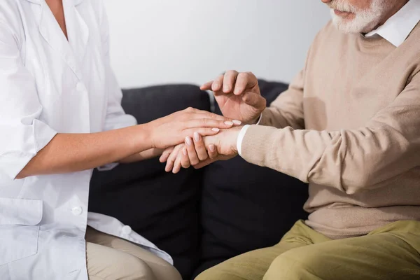 Cropped view of senior man and social worker holding hands while sitting on sofa at home — Stock Photo