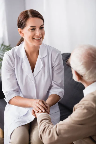 Lächelnde Krankenschwester hält die Hand eines betagten Mannes, während er zu Hause auf dem Sofa sitzt — Stockfoto