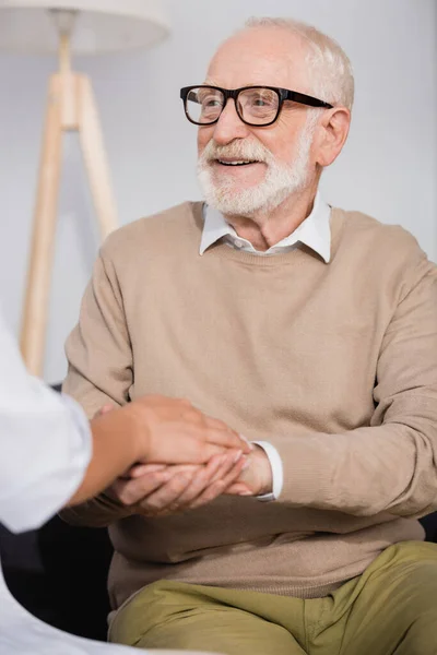 Sorridente homem idoso de mãos dadas com a enfermeira social em casa em primeiro plano desfocado — Fotografia de Stock