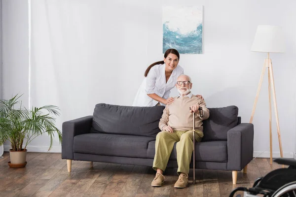 Улыбающаяся медсестра обнимает пожилого мужчину сидящего на диване с тростью — стоковое фото