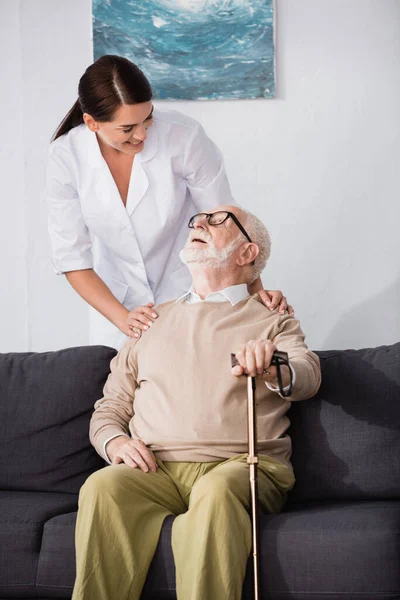 Homme âgé souriant assis sur le canapé avec bâton de marche et regardant travailleur social — Photo de stock