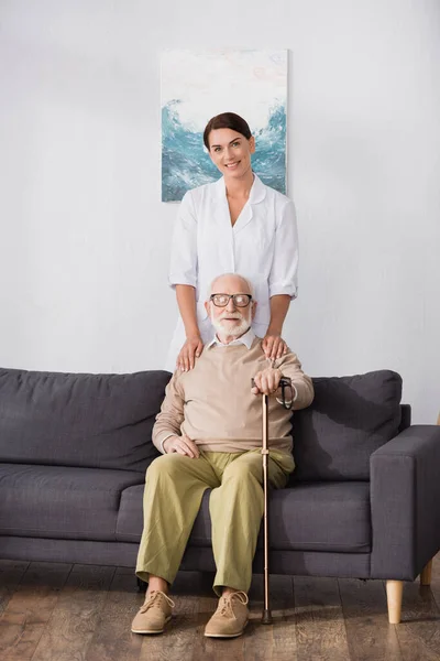 Веселый социальный работник трогает плечи пожилого человека, сидящего на диване с тростью — стоковое фото