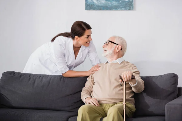 Enfermera geriátrica sonriente hablando con un anciano sentado en un sofá en casa - foto de stock
