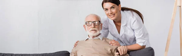 Геріатрична медсестра обіймає плечі літнього чоловіка, дивлячись на камеру, банер — стокове фото