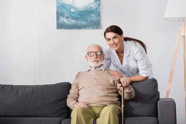 Assistente sociale sorridente che abbraccia l'uomo anziano seduto sul divano di casa — Foto stock