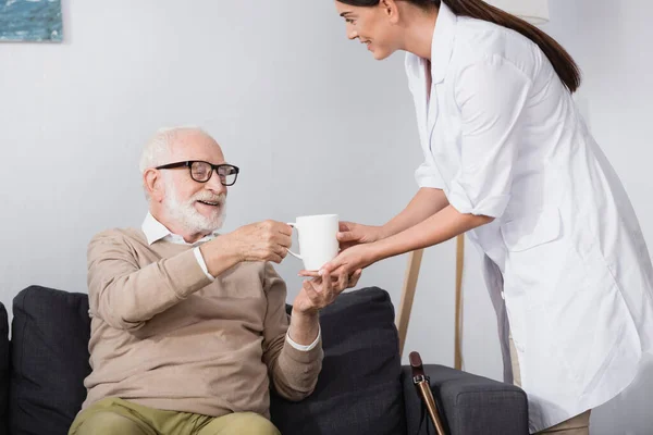 Старческая медсестра, дающая чашку чая пожилому мужчине, сидящему на диване — стоковое фото
