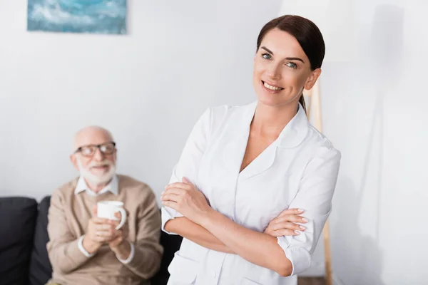 Allegro assistente sociale sorridente alla macchina fotografica mentre in piedi con le braccia incrociate vicino all'uomo anziano in possesso di una tazza di tè — Foto stock