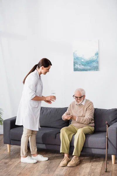 Elderly man sitting on sofa and holding pills while medical assistant glass of water — Stock Photo