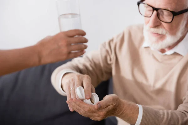 Anciano en anteojos tomando pastillas cerca de la enfermera dando vaso de agua en primer plano borrosa - foto de stock