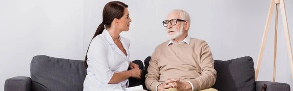 Geriatric nurse talking to aged man while sitting on sofa at home, banner — Stock Photo