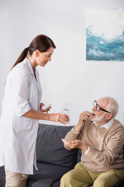 Bruna infermiera sociale che dà un bicchiere d'acqua e pillole all'uomo anziano a casa — Foto stock