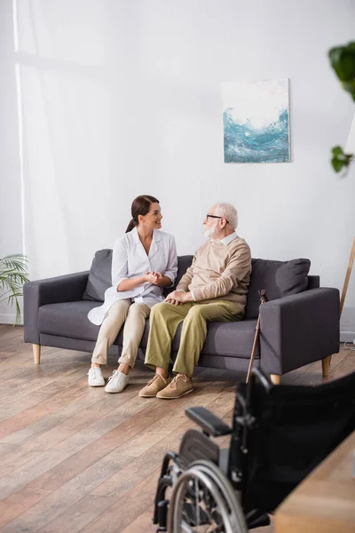 Travailleur social parler à un homme âgé assis sur le canapé à la maison avec fauteuil roulant au premier plan flou — Photo de stock