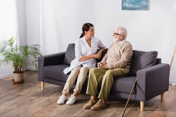 Brünette Sozialarbeiterin und alter Mann reden, während sie zu Hause auf dem Sofa sitzen — Stockfoto