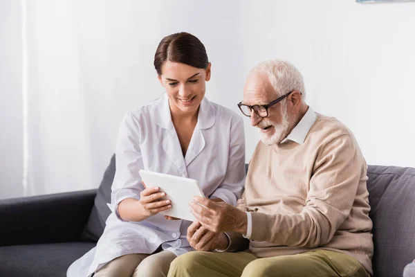 Brunette social worker showing digital tablet to smiling aged man at home — Stock Photo
