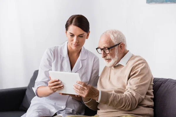 Enfermera geriátrica morena y hombre de edad avanzada usando tableta digital juntos - foto de stock