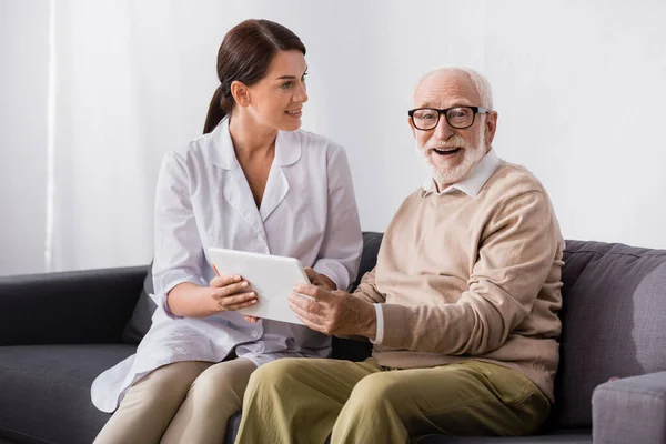Aufgeregter Senior blickt in Kamera, während er digitales Tablet in der Nähe der Altenpflegerin hält — Stockfoto