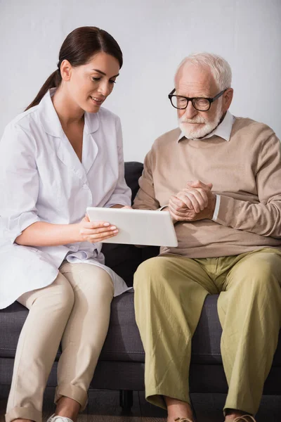 Brünette Altenpflegerin zeigt älteren Mann, der mit geballten Händen sitzt, ein digitales Tablet — Stockfoto