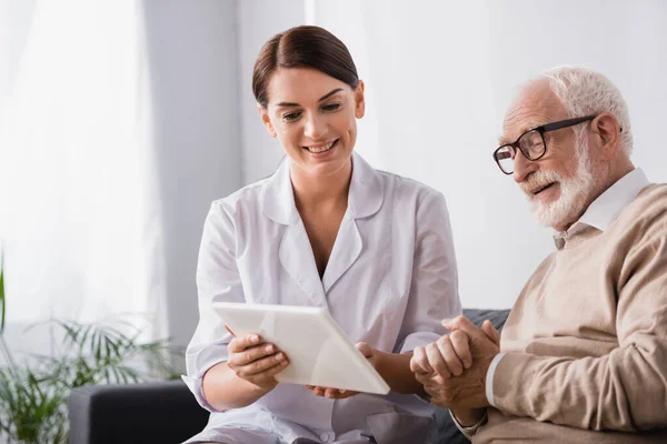 Smiling social worker showing dgital tablet to senior man sitting with clenched hands — Stock Photo