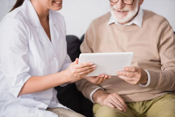 Vista recortada de la enfermera geriátrica mostrando tableta digital al hombre anciano sobre fondo borroso - foto de stock