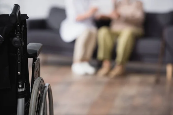 Assistant médical et homme âgé assis sur le canapé à la maison près du fauteuil roulant au premier plan flou — Photo de stock