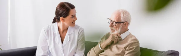Geriatric nurse and aged man talking while sitting on sofa at home, banner — Stock Photo
